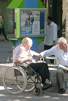 Photo of two older men, one in a wheelchair, sitting in a public, outdoor, urban space